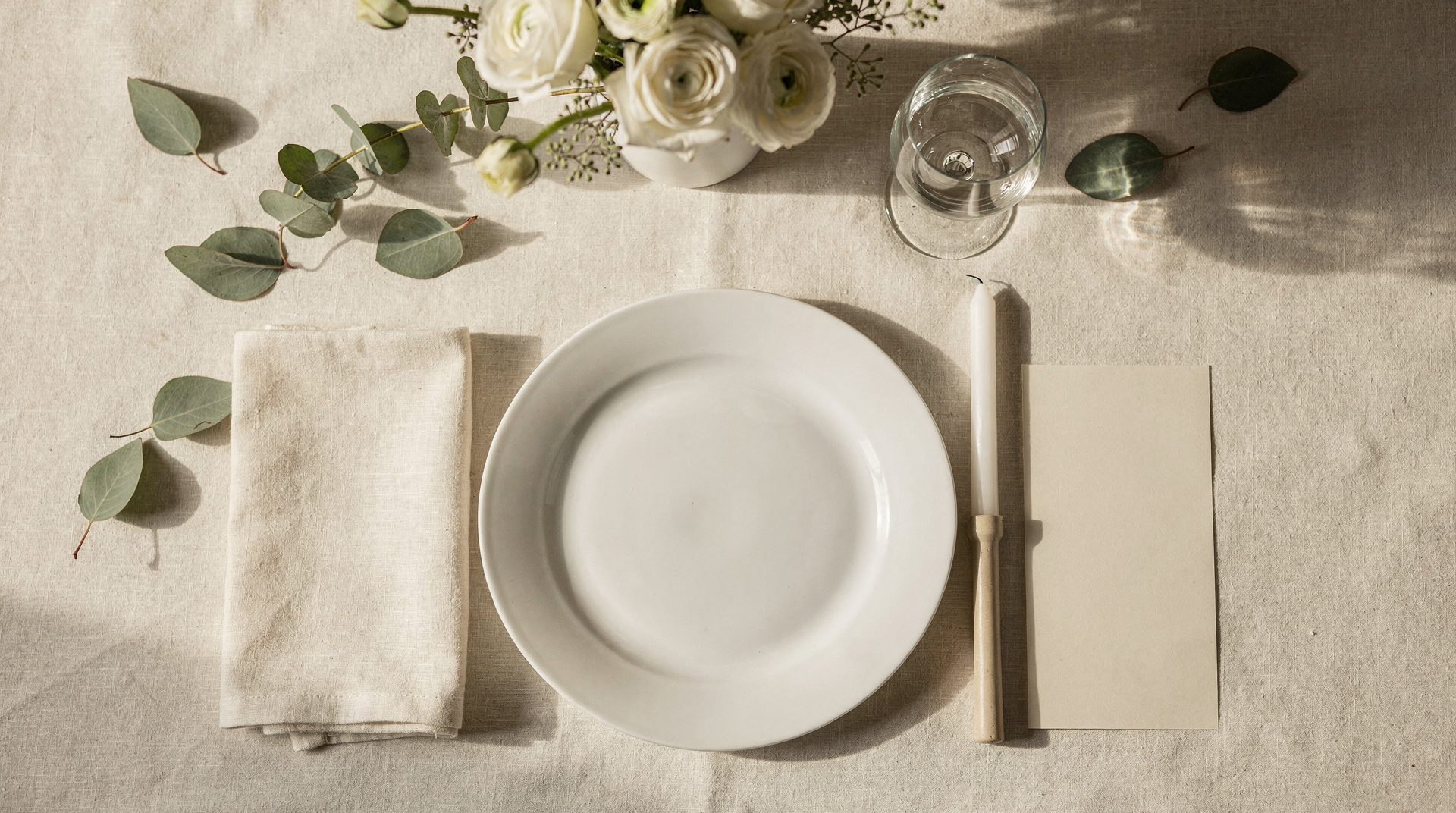 Flatlay linen krem: piring kosong elegan, gelas, serbet lipat, bunga meja rendah, lilin putih, kartu menu kosong, dan daun eucalyptus